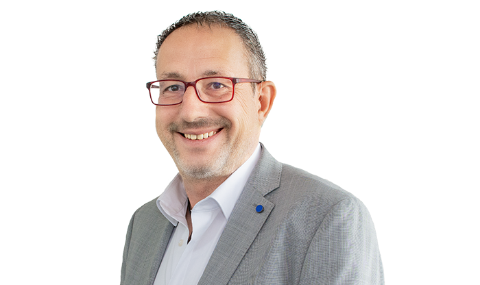 Portrait eines Mitarbeiters mit kurzem Haar, einer Brille, einem weißen Hemd und einem hellen Sakko. 